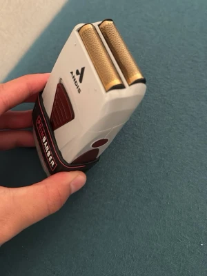 Andis rakapparat med PrimBarber grepp - Elektrisk rakapparat från Andis med dubbla guldiga foliehuvuden och ergonomiskt grepp från PrimBarber. Vit och mörkröd design med robust plastmaterial och detaljer i gummi för extra komfort. Perfekt för precision och smidig rakning.