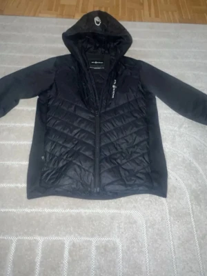 Sail racing jacka - Snygg svart pufferjacka från All Szn med huva och dragkedja framtill. Jackan har quiltad design på framsidan och logga på både bröstet och huvan. Tillverkad i ett glansigt material på kroppen och mjukare tyg på ärmarna. Perfekt för kalla vinterdagar.