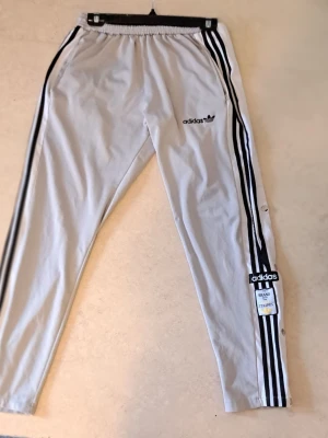 Adidas grå träningsbyxor med knappar - Grå träningsbyxor från Adidas med svarta klassiska tre ränder längs sidorna och tryckt logga på låret. Byxorna har knappar längs benen för att kunna öppnas upp och elastisk midja. Perfekta för sport eller chill.