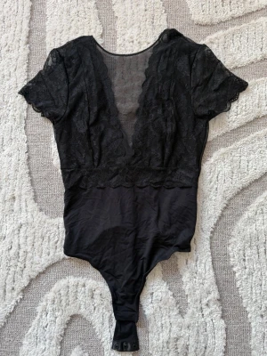 Svart spetsbody med djup V-ringning - Snygg svart body med korta ärmar och djup V-ringning framtill. Överdelen är i transparent spets med blommönster och nederdelen i mjukt tyg. Knäppning i grenen. Helt oanvänd.