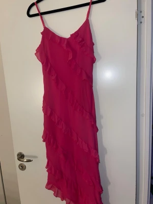 Rosa volangklänning med smala axelband - Snygg rosa klänning med smala axelband och volangdetaljer som går diagonalt över hela klänningen. Lätt och luftig modell som ger en feminin look. Perfekt för varma dagar eller när du vill sticka ut lite extra.