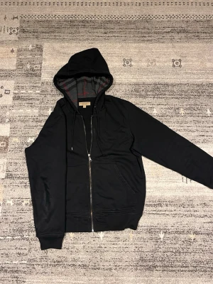 Äkta Svart Burberry hoodie - Säljer nu en snygg, stilren svart hoodie från Burberry med dragkedja framtill och två fickor. Insidan av huvan har klassiskt Burberry-mönster i grått, svart och rött. Tillverkad i mjukt bomullsmaterial och har justerbara snören i huvan. Perfekt för en chill och stilren look. Perfekt nu för våren. Hoodien passar M. Obs… den är äkta och kvitto går att lösa. Kan tänka mig gå ner nån hundralapp vid snabbaffär. 