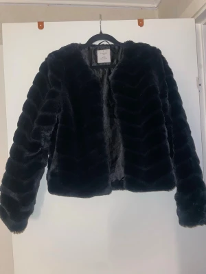 Svart kort pälsjacka från Linette - Snygg svart fuskpälsjacka från Linette med kortare passform och långa ärmar. Jackan har ett stilrent, mönstrat pälsmaterial och är öppen framtill utan synliga knappar. Perfekt för att lyfta din vinterstil.