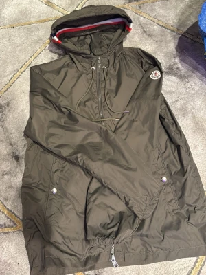 Moncler mörkgrön vindjacka med huva - Snygg mörkgrön vindjacka från Moncler med huva och dragsko. Jackan har två fickor med tryckknappar och klassisk Moncler-logga på ärmen. Lätt material som passar perfekt för blåsiga dagar. Röd, vit och blå detalj i huvkanten.