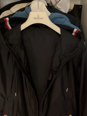 Svart vindjacka från Moncler - Snygg svart vindjacka från Moncler med dragkedja, huva och klassiska Moncler-loggan på ärmen. Jackan har diskreta röda och vita detaljer på huvan och är tillverkad i ett lätt, vattenavvisande material. Perfekt för dig som vill ha en stilren och sportig look.