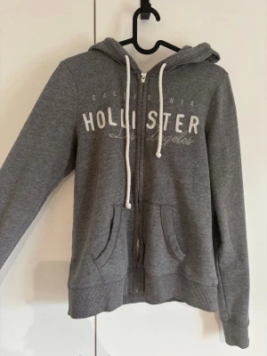 Grå zip-hoodie från Hollister🫶🏻 - Snygg grå hoodie från Hollister med dragkedja, vita snören och tryck på bröstet där det står 'Hollister Los Angeles'. Mjuk insida och två fickor framtill. Perfekt för en avslappnad och cool stil. Har ett litet hål i luvan (se bild fem)🤍