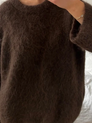 Mörkbrun stickad tröja - Mysig mörkbrun stickad tröja med fluffig yta och rund hals. Tröjan har en avslappnad passform och långa ärmar, perfekt för kyliga dagar. Materialet är mohair eller en liknande ullblandning som ger en extra mjuk känsla.