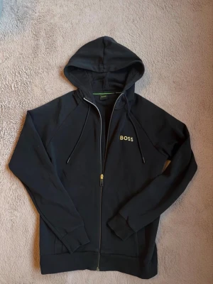 Svart zip-hoodie från hugo boss - Snygg svart hoodie från hugo boss med dragkedja framtill och justerbar huva med snören. Klassisk design med BOSS-logga i gult på bröstet. Tillverkad i mjukt bomullsmaterial och har ribbade muddar vid ärmslut och nederkant. Perfekt för en chill och stilren look. Det står storlek XS men passar även S