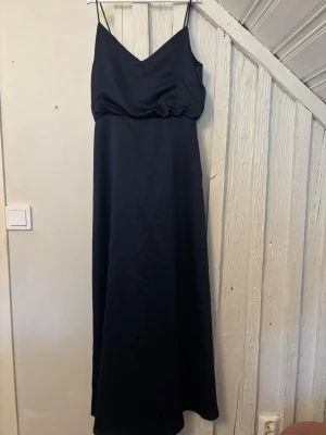 Marinblå långklänning med smala band - Elegant marinblå långklänning med smala axelband och v-ringad urringning både fram och bak. Klänningen har en enkel, stilren design och är lätt utsvängd nertill. Perfekt för dig som gillar minimalistisk stil.