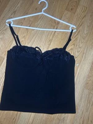 Topp - Svart korsettopp från Gina med tunna axelband, spetsdetaljer upptill och snörning framtill. Toppen har en dragkedja i ryggen och en figurnära passform. Perfekt för att lyfta vilken outfit som helst. Helt oanvänd med prislapp kvar