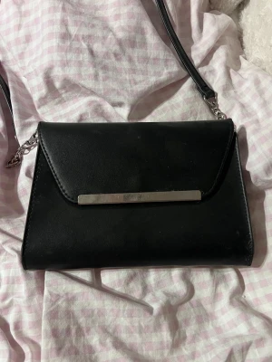 Svart kuvertväska med kedjedetalj - Stilren svart kuvertväska i slätt konstläder med lång axelrem och kedjedetaljer. Väskan har ett elegant silverfärgat metallspänne på framsidan och en enkel, rektangulär form. Perfekt för dig som gillar minimalistisk och modern design.