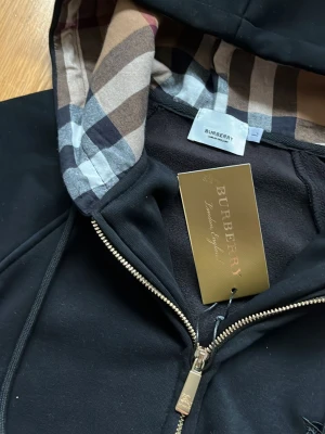 Svart hoodie från Burberry med dragkedja - Svart hoodie från Burberry med klassiskt rutigt foder i huvan och guldfärgad dragkedja. Broderad Burberry-logga på bröstet och mjukt bomullsmaterial. Perfekt för dig som vill ha en stilren och lyxig look.