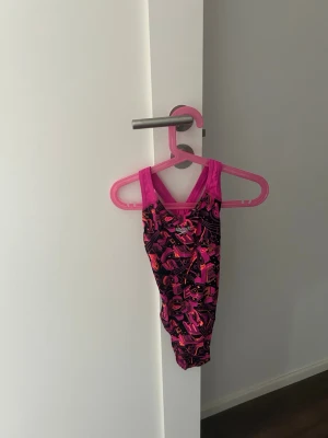 Färgglad baddräkt från Speedo - Snygg baddräkt från Speedo med färgstarkt grafiskt mönster i rosa, lila och orange toner. Axelbanden är breda och rosa, vilket ger en sportig look. Perfekt för simträning eller strandhäng. Materialet är troligtvis polyester eller liknande snabbtorkande tyg.