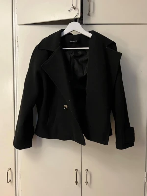Kort svart kappa  - Stilren svart kappa med kortare passform och breda slag. Kappan har en enkelknäppning framtill och är helfodrad. Perfekt för dig som vill ha en klassisk men trendig look till hösten eller vintern.
