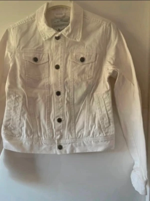 Vit dam jeansjacka med svarta knappar - Snygg vit jeansjacka med klassisk krage och svarta knappar framtill. Två bröstfickor med lock och knapp samt två sidofickor. Rak passform och långa ärmar. Perfekt att slänga över en t-shirt för en cool look. Storlek 34, men upplevs som 36. Används 2 gånger.