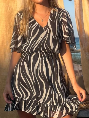 Zebramönstrad kort klänning - Snygg kort klänning med zebramönster i svart och vitt. Klänningen har v-ringning, volangkant nertill och löst sittande korta ärmar. Perfekt för sommarens alla tillfällen och ger en trendig look.