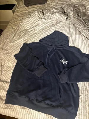 Mörkblå hoodie från Stüssy - Säljer en mörkblå hoodie från Stüssy med klassisk logga tryckt i vitt på bröstet. Hoodien har huva med dragsko och en stor magficka. Tillverkad i mjukt bomullsmaterial och har ribbade muddar vid ärmslut och nederkant. Perfekt för en avslappnad streetwear-look.