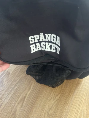 Basketväska - Svart ryggsäck Spånga Basket - Säljer en svart ryggsäck med vit text 'Spånga Basket' på locket. Ryggsäcken har ett stort nätfack framtill, perfekt för att bära med sig träningskläder eller skor. Tillverkad i slitstarkt tyg och mesh, sportig och praktisk design.