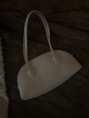 Vit bågformad handväska - Stilren vit handväska med bågformad design och två långa handtag. Väskan har en minimalistisk look och är tillverkad i slätt material som ger en modern känsla.