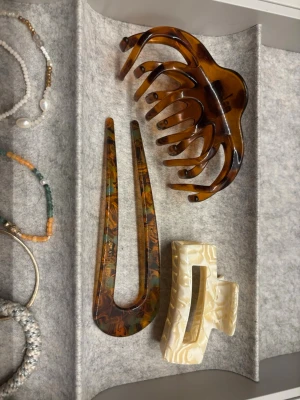 Set med hårklämma och hårsticka - Säljer ett set med tre håraccessoarer: en stor brun hårklämma i sköldpaddsmönster, en hårsticka i brunt och grönt mönster samt en rektangulär hårklämma i pärlemorvit plast. Perfekta för att skapa snygga frisyrer och hålla håret på plats.