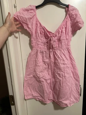 Rosa kort puffärmad klänning storlek: 44 - Söt rosa kortklänning med puffärmar och v-ringning. Klänningen har snörning framtill och resår i midjan för en bekväm passform. Perfekt för varma dagar och har en luftig känsla.