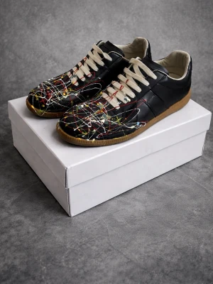 Maison margiela - Unika svarta sneakers med färgstänk i vitt, rött, blått och gult. Skorna har vita snören, låg profil och en klassisk gummisula. Ovandelen är i läder och designen ger en konstnärlig och modern vibe. Perfekta för dig som vill sticka ut med din stil.