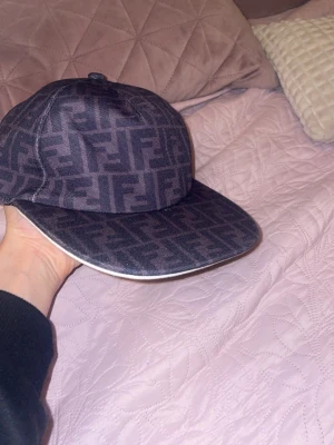 Fendi keps med FF-mönster - Snygg keps från Fendi i mörkgrått med det klassiska FF-logomönstret över hela kepsen. Kepsen har böjd skärm med vit kant och är tillverkad i ett tygmaterial som känns både lyxigt och street. Perfekt för dig som vill sticka ut med designeraccessoar.