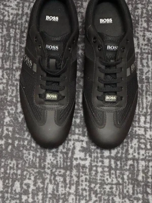 Svarta sneakers från Hugo BOSS - Stilrena svarta sneakers från BOSS med diskret logga på sidan och tungan. Skorna har en kombination av läder och textilmaterial, svarta skosnören och låg profil. Perfekta för en clean och modern look.