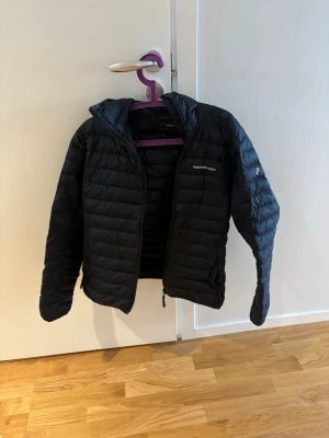 Svart pufferjacka från Peak Performance - Snygg svart pufferjacka från Peak Performance med huva och diskret logga på bröstet. Jackan har quiltad design och dragkedja framtill. Perfekt för kalla dagar när du vill hålla värmen och ändå se stilren ut. Storlek M 