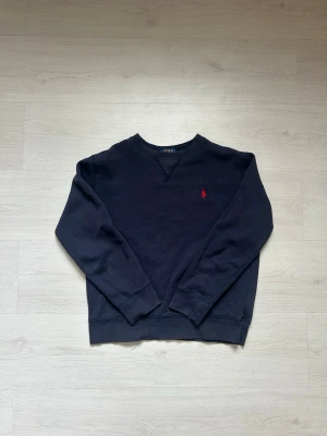 Mörkblå tröja från Polo Ralph Lauren - Det här är en marinblå Polo Ralph Lauren-tröja. Den har varumärkets ikoniska polospelarlogotyp i rött, broderad på bröstet. Tröjan har en klassisk rund halsringning och en avslappnad passform, vilket gör den perfekt för fritidskläder. Tyget verkar vara en mjuk, troligen bomullsblandning, lämplig för kallare väder. Den övergripande stilen är tidlös, med fokus på komfort och varumärkesigenkänning.