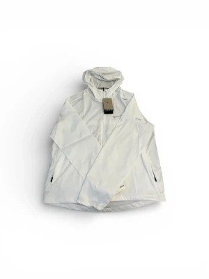 Vit vindjacka från Nike med huva - Storlek (M) Snygg vit vindjacka från Nike med dragkedja framtill, två sidofickor med dragkedja och justerbar huva. Jackan har diskret Nike-logga på bröstet och är tillverkad i lätt, vindtätt material. Perfekt för blåsiga dagar och sportiga outfits.