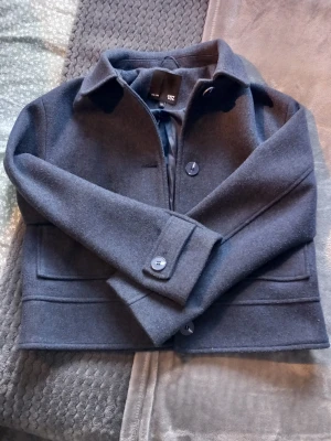 Mörkblå ullcardigan med fickor - Snygg mörkblå cardiganjacka från Lager 157 i ullblandning. Jackan har stora knappar framtill, två rejäla fickor och klassisk krage. Rak och boxig passform som ger en trendig look. Perfekt för lager-på-lager-stil under kyliga dagar. Tyget har dock gått upp i båda ärmarna, men lätt att laga. 