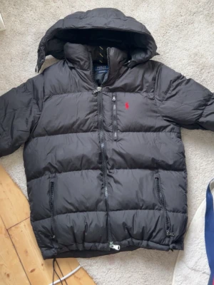 Svart pufferjacka från Polo Ralph Lauren - Svart pufferjacka från Polo Ralph Lauren med huva och dragkedja framtill. Jackan har en röd broderad logga på bröstet, flera fickor med dragkedja och breda quiltade sektioner. Perfekt för kalla dagar och riktigt skön att bära.