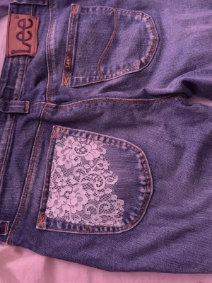 Lee jeans med spetsdetaljer - Unika blå Lee jeans med utsvängda ben och snygga vita spetsdetaljer på bakficka och benslut. Klassisk femficksmodell med cool vintagekänsla och kontrastsömmar. Perfekta för dig som vill sticka ut med en personlig stil.