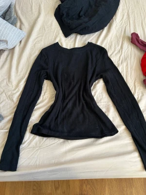 Svart ribbad långärmad topp - Svart långärmad topp med ribbad struktur och rund halsringning. Modellen är figurnära och har en enkel, stilren design som passar till många olika outfits. Perfekt basplagg för garderoben.