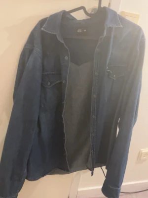 Blå jeansskjorta med knappar - Snygg blå jeansskjorta från H&M i klassisk modell med långa ärmar och tryckknappar framtill. Skjortan har två bröstfickor med lock och är gjord i mjuk bomull. Perfekt att bära öppen över en t-shirt eller stängd för en mer stilren look.