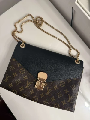 Monogram axelremsväska - Snygg handväska med klassiskt monogrammönster i brunt och guld. Väskan har svart lock i läder, guldfärgad kedjerem och elegant låsdetalj framtill. Röd på insidan❤️