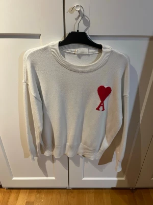 Vit stickad tröja från Ami Paris - Vit stickad tröja från Ami Paris med rund halsringning och ribbade muddar. Tröjan har en röd hjärtlogga med bokstaven 'A' broderad på bröstet. Perfekt för dig som gillar stilrena plagg med en twist.