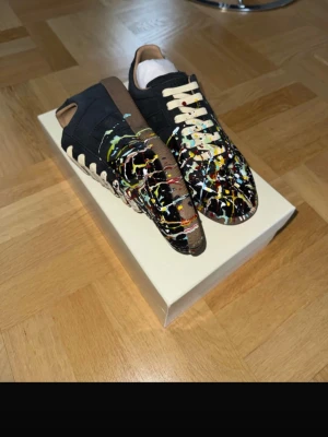 Maison Margiela sneakers med färgstänk - Unika sneakers från Maison Margiela i svart läder och mocka med beige snörning och färgstänk i vitt, gult, rött och blått. Låg modell med klassisk siluett och bruna detaljer vid häl och plös. Perfekta för dig som vill sticka ut med din stil.