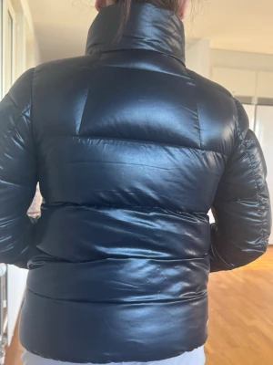 Svart pufferjacka med hög krage - Säljer en svart pufferjacka med glansig finish och hög krage. Jackan har breda, vadderade paneler och dragkedja framtill. Perfekt för kalla dagar när du vill hålla värmen och ändå se stilren ut. Köpte den som storlek 16 men skulle säga att det motsvarar xs/s.