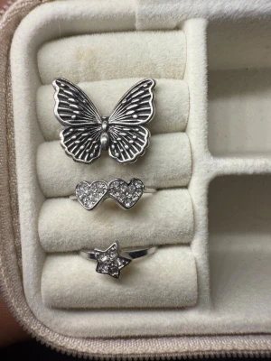 Silver ringar  - Säljer tre snygga silvriga ringar: en med en stor fjäril, en med två hjärtan täckta av glittrande stenar och en med en stjärna dekorerad med små stenar. Perfekta accessoarer för att lyfta din stil. 39kr/st🩷🩷
