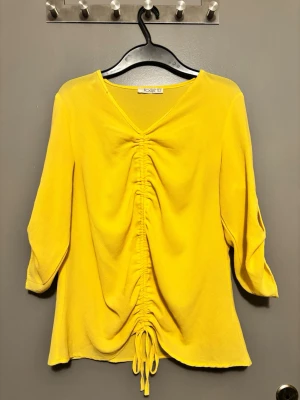 Gul blus med rynk framtill köpt utomlands  - Snygg gul blus från focus med v-ringning och rynkdetalj framtill som kan justeras med knytband. Blusen har trekvartsärm med knappdetalj och är tillverkad i ett lätt, strukturerat tyg som ger en luftig känsla.