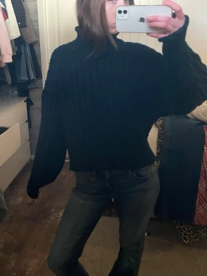 Svart ribbstickad tröja med hög krage - En svart, ribbstickad tröja med hög krage och långa ärmar. Tröjan har en avslappnad passform och är perfekt att matcha med jeans. Materialet ser ut att vara mjukt och varmt, idealiskt för kyliga dagar.