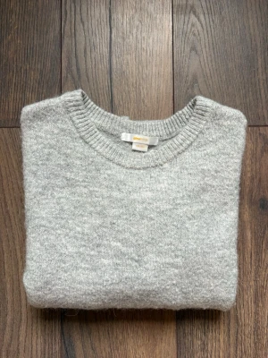 Grå stickad t-shirt från Gina Tricot - Säljer en stilren grå stickad t-shirt från Gina Tricot. Tröjan har korta ärmar, rund halsringning och är gjord i ett mjukt, melerat material som känns skönt mot huden. Perfekt för dig som gillar enkel och clean stil.