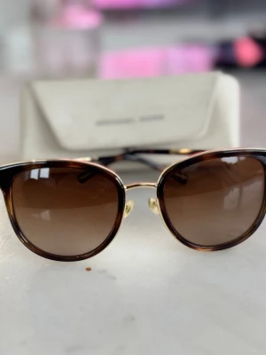 Bruna solglasögon Michael Kors - Snygga solglasögon från Michael Kors med bruna glas och guldfärgade detaljer. Bågarna har en klassisk rundad form och är tillverkade i en kombination av metall och syntet. Perfekta för att lyfta din stil under soliga dagar.
