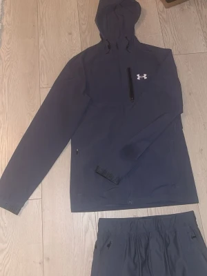 Under Armour mörkgrå  - Snyggt mörkgrått sett från Under Armour med matchande hoodie och byxor. Hoodien har dragkedja, justerbar huva och bröstficka med logga. Byxorna har resår i midjan och dragkedjor vid fickorna samt Under Armour-logga på benet. Perfekt för träning eller chill.