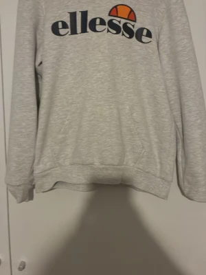 Grå sweatshirt från Ellesse - Snygg grå sweatshirt från Ellesse med stor logga i marinblått och orange framtill. Tröjan har rund hals, långa ärmar och ribbade muddar vid ärmslut och nederkant. Perfekt för en avslappnad och sportig stil.