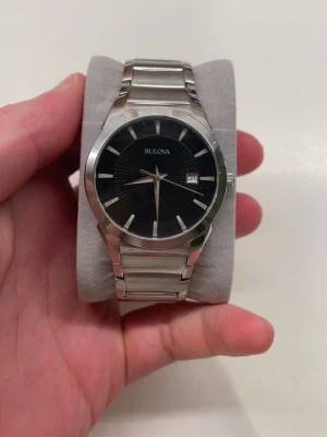 Bulova silverfärgad klocka - Stilren klocka från Bulova med svart urtavla och datumvisning. Klockan har ett klassiskt armband i silverfärgat metall och minimalistiska indexmarkeringar. Perfekt accessoar för dig som gillar en tidlös och elegant look. Några repor på glaset. 