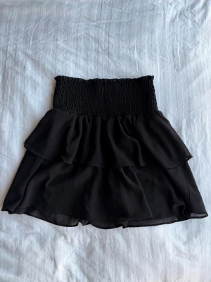 Svart volangkjol med resårmidja - Säljer en svart kortkjol med bred smockresår i midjan och dubbla volanger. Kjolen är luftig och har en flowy känsla, perfekt till sommaren. Materialet känns som lätt polyester eller syntet, vilket gör den bekväm och enkel att bära.