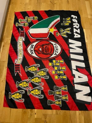 AC Milan supporterflagga - Stor supporterflagga för AC Milan i rött och svart med klubbens emblem, texten 'Forza Milan' och flera troféer tryckta. Flaggan har även italienska färger och guldstjärna. Perfekt för att visa ditt stöd på matcher eller hemma.